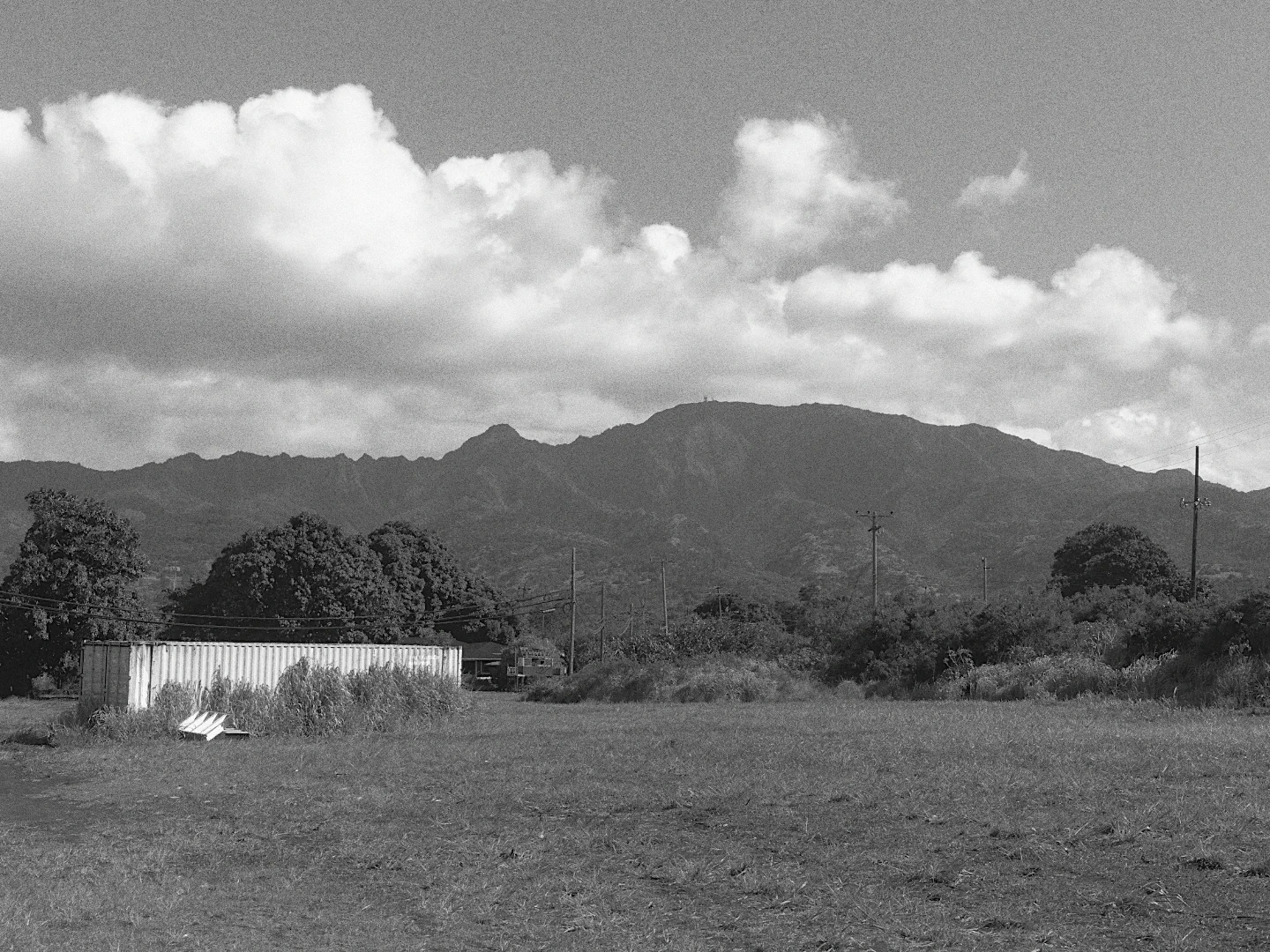 A field below a mountain ridge under white clouds, in black and white with Ilford HP5 Plus 400.